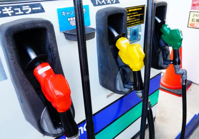 ガソリンスタンドの給油ノズルと、レギュラー・ハイオク・軽油の価格ラベルが並んでいる様子。