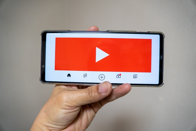 スマートフォンで動画を再生するイメージ。赤い再生ボタンが映し出された画面を手に持っている様子です。